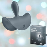 A gray curved plug is placed against a soft, multicolored bokeh background. To the right, a box labeled "Satisfyer Booty Absolute Beginners 2 Vibrating Anal Plug" provides information on its functionality for those new to anal play and emphasizes its vibration feature for the ultimate Booty Bliss.